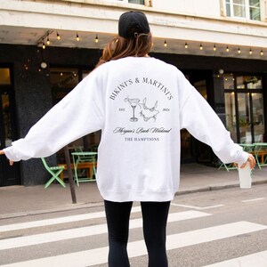 以下が含まれることがあります： 「Bikini's & Martinis Est. 2024 Morgan's Bach Weekend The Hamptons」という黒い文字が書かれた白いスウェットシャツ。スウェットシャツには、マティーニグラスとビキニの白黒イラストが描かれている。