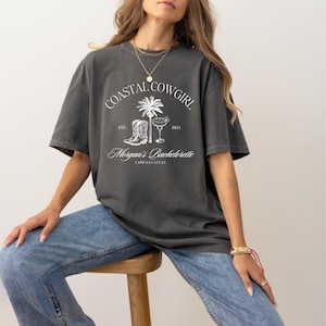 Puede incluir: Una camiseta gris con un dise&ntilde;o gr&aacute;fico blanco que dice "Coastal Cowgirl Est. 2021 Morgan's Bachelorette Cabo San Lucas". El gr&aacute;fico tambi&eacute;n incluye una palmera, una copa de margarita y botas de vaquero.
