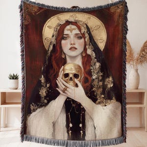 May include: Woven tapestry throw blanket featuring a Renaissance-style portrait of a woman with red hair, holding a golden skull. The artwork has a dark red background with gold accents and a fringed edge. The woman is wearing a black veil.