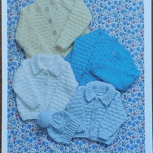 Może przedstawiać: Cztery dzianinowe sweterki dla niemowląt w pastelowych kolorach: żółtym, niebieskim, białym i jasnoniebieskim. Sweterki mają zapięcia na guziki i są prezentowane na tle kwiatowym. Do jasnoniebieskiego sweterka dołączona jest pasująca czapka.