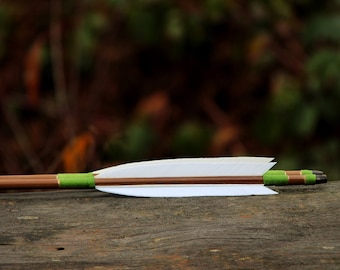 May include: A white fletched arrow with green and brown accents rests on a weathered wooden surface.