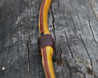 May include: A wooden bow with a red and yellow stain. The bow has a leather wrap around the handle.