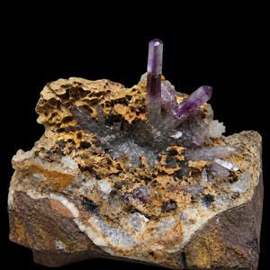 May include: A natural mineral specimen featuring translucent purple amethyst crystal points emerging from a porous, textured tan and brown rock matrix. The cluster displays varied crystalline formations with deep violet tips and clear quartz accents.