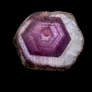 Puede incluir: Una piedra preciosa en bruto de forma hexagonal que presenta un centro translúcido de color púrpura intenso con patrones geométricos concéntricos. Los bordes exteriores son opacos, texturizados y de color blanco a gris claro.