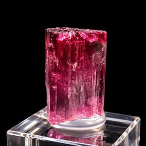 Pode incluir: Um cristal de turmalina rosa profundo e vibrante com uma estrutura colunar estriada verticalmente e uma borda superior translúcida e irregular. A pedra preciosa está montada numa base de exibição quadrada de acrílico transparente.