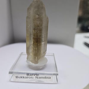 Puede incluir: Cristal de barita translúcido de color marrón claro de Bukkos, Namibia, expuesto sobre un soporte acrílico transparente. El cristal tiene forma columnar con una parte superior puntiaguda. El soporte tiene una etiqueta con el nombre y el origen del cristal.