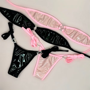 May include: Two pairs of bikini bottoms, one black and one pink. Both pairs have a shiny, wet look finish and feature a heart-shaped rhinestone embellishment.