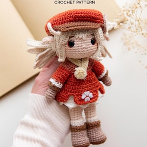 May include: A Klee crochet doll featuring a beige face, blonde hair, and a red hat. The doll wears a red dress with a white four-leaf clover emblem, a white collar with a small pom-pom, and brown boots. The text Klee CROCHET PATTERN is visible above.