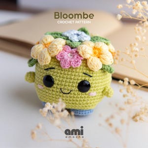 May include: A light green crocheted amigurumi figure with a smiling face, black eyes, and pink cheeks. The top is adorned with yellow, pink, and light blue crocheted flowers and green leaves. The text Bloombe CROCHET PATTERN is visible above.