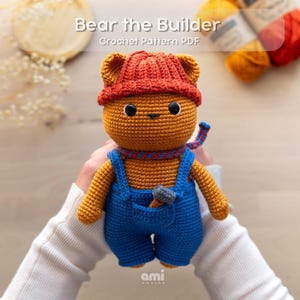 May include: A brown crocheted bear toy wearing a red beanie, a blue and red patterned scarf, and blue overalls. A small grey hammer is tucked into the front pocket of the overalls. The text Bear the Builder Crochet Pattern PDF is visible.