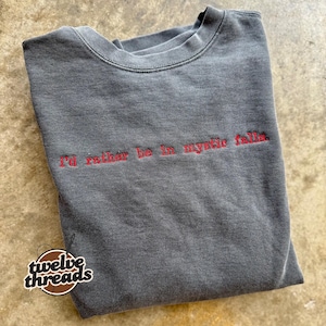 May include: A charcoal grey crewneck sweatshirt featuring the red embroidered text 'i'd rather be in mystic falls.' across the chest. The garment has a faded, washed-out appearance and a round neckline with ribbed trim detailing.