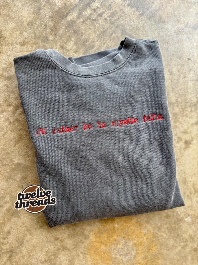 May include: A charcoal grey crewneck sweatshirt featuring the red embroidered text 'i'd rather be in mystic falls.' across the chest. The garment has a faded, washed-out appearance and a round neckline with ribbed trim detailing.