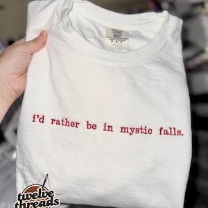 May include: A white short-sleeved t-shirt featuring the embroidered red text "i'd rather be in mystic falls." centered on the chest. The garment has a crew neckline and a visible interior brand label at the collar area.