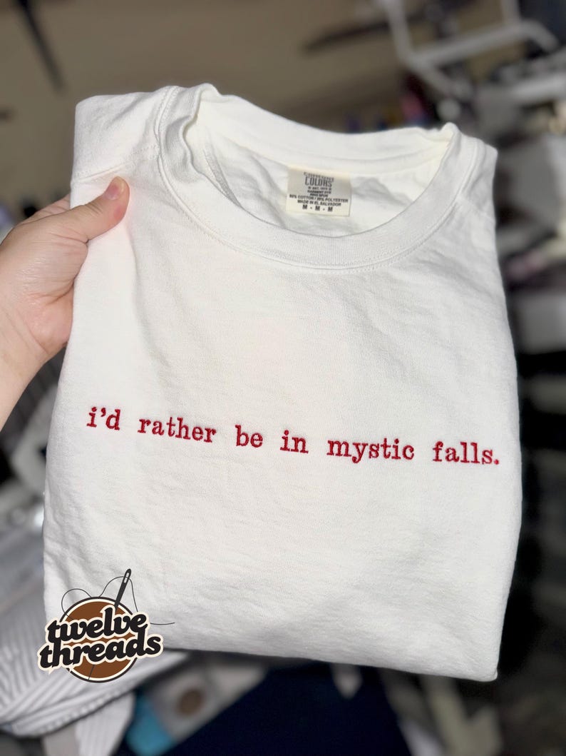May include: A white short-sleeved t-shirt featuring the embroidered red text "i'd rather be in mystic falls." centered on the chest. The garment has a crew neckline and a visible interior brand label at the collar area.