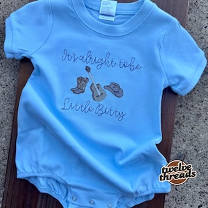May include: Light blue baby romper with short sleeves. The romper features embroidered images of cowboy boots, a guitar, and a cowboy hat, along with the text "It's alright to be Little Bitty."