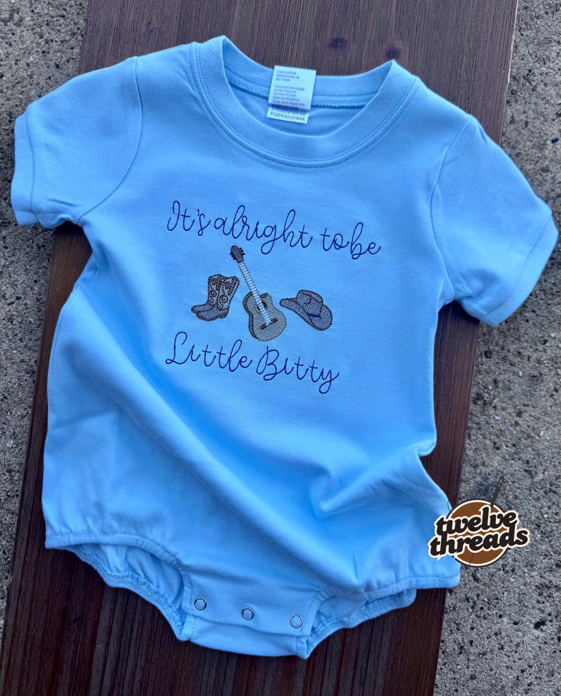 May include: Light blue baby romper with short sleeves. The romper features embroidered images of cowboy boots, a guitar, and a cowboy hat, along with the text "It's alright to be Little Bitty."