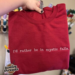 May include: A dark red crewneck sweatshirt featuring the white embroidered text "i'd rather be in mystic falls." centered on the chest. The garment has a ribbed neckline and a standard fit, showcasing a clean and simple design aesthetic.