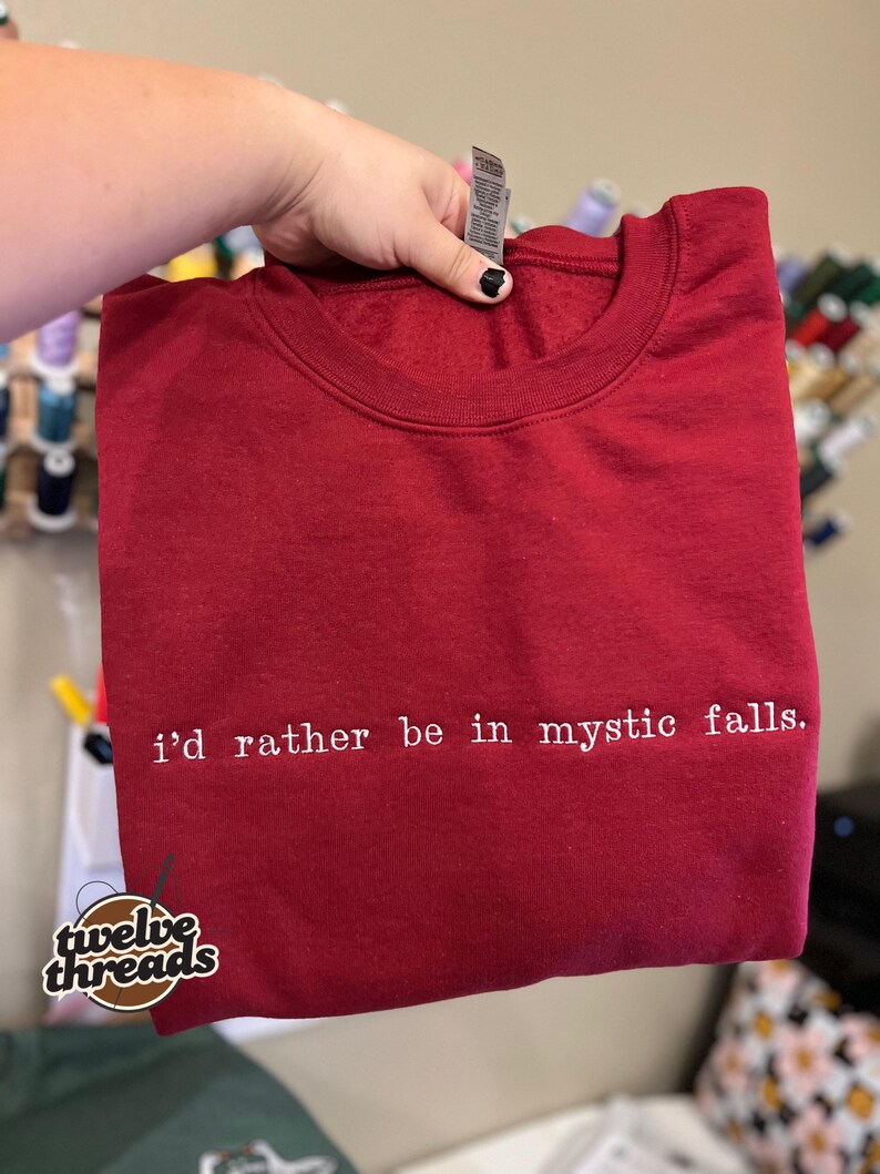 May include: A dark red crewneck sweatshirt featuring the white embroidered text "i'd rather be in mystic falls." centered on the chest. The garment has a ribbed neckline and a standard fit, showcasing a clean and simple design aesthetic.