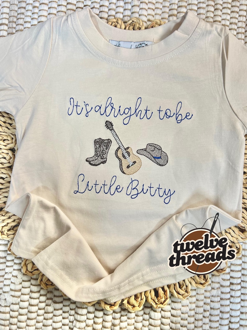 May include: Cream-colored t-shirt with embroidered design. The design includes a guitar, cowboy boots, and a hat, with the text "It's alright to be Little Bitty" in blue script. The shirt has a round neckline and short sleeves.