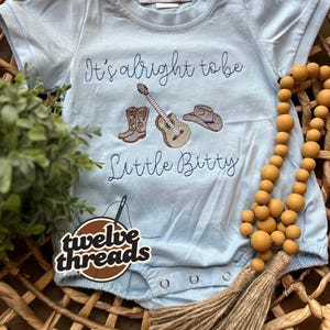 May include: Light blue baby bodysuit with embroidered text "It's alright to be Little Butty" and cowboy-themed graphics. The bodysuit features a guitar, cowboy boots, and a cowboy hat. The item is displayed in a woven basket with wooden beads.