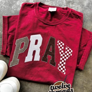 Puede incluir: Una camiseta roja con la palabra "PRAY" aplicada en la parte delantera. Las letras presentan una mezcla de patrones, incluyendo cuadros vichy y rayas. Zapatillas blancas visibles en la esquina inferior izquierda. El logotipo de la marca "twelve threads" está en la esquina inferior derecha.