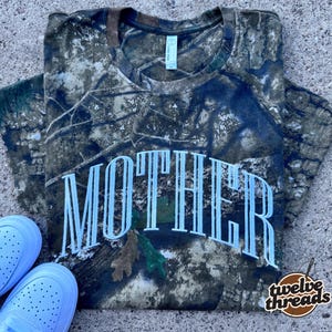 May include: Camouflage-patterned t-shirt with the word "MOTHER" in large white letters across the front. The shirt is paired with white sneakers. The brand "twelve threads" is printed in the lower right corner.