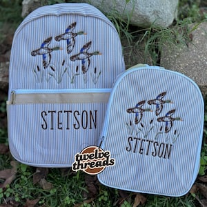 Op de afbeelding: Een lichtgrijze en witte rugzak met krijtstreep en een bijpassende lunchtas. Beide items zijn voorzien van geborduurde wilde eenden die over riet vliegen en de naam STETSON in bruin garen. De rugzak heeft een horizontale beige accentstrook.