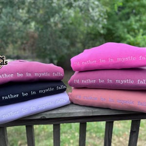 May include: A stack of six folded sweatshirts in various shades of pink, purple, and black. Each sweatshirt features the embroidered white text "i'd rather be in mystic falls." The garments are arranged in two columns on a wooden railing.