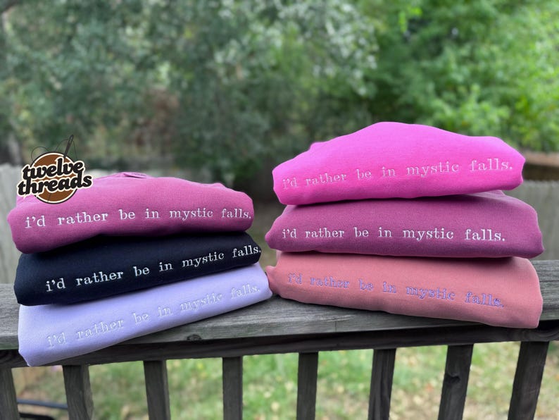 May include: A stack of six folded sweatshirts in various shades of pink, purple, and black. Each sweatshirt features the embroidered white text "i'd rather be in mystic falls." The garments are arranged in two columns on a wooden railing.