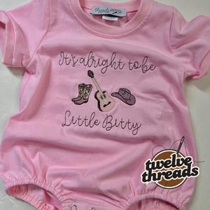 May include: Pink baby romper with the embroidered text "It's alright to be Little Bitty" and images of a cowboy boot, guitar, and cowboy hat. The romper has short sleeves and snaps at the bottom.