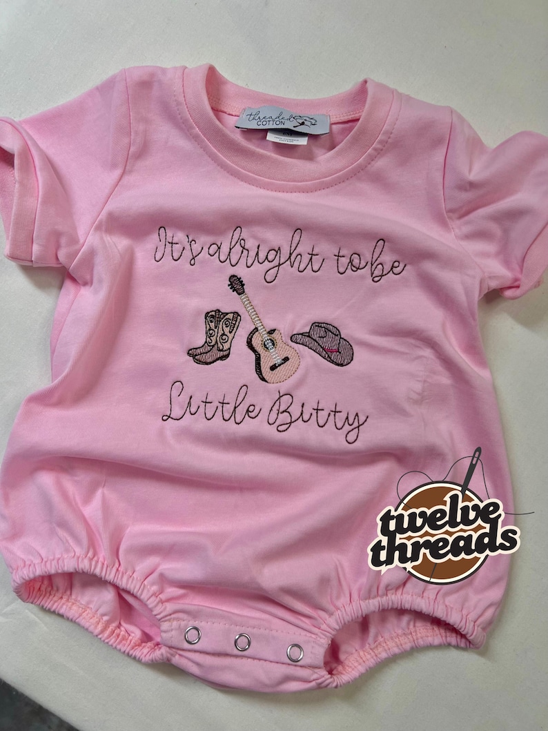 May include: Pink baby romper with the embroidered text "It's alright to be Little Bitty" and images of a cowboy boot, guitar, and cowboy hat. The romper has short sleeves and snaps at the bottom.