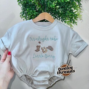 May include: A beige baby bodysuit with rolled sleeves, featuring embroidered text and graphics. The text reads "It's alright to be Little Bitty" above a guitar, cowboy boots, and a hat. The brand logo "twelve threads" is on the right.