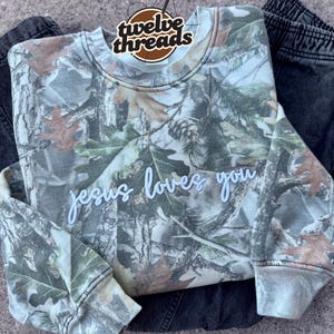 May include: A camouflage-patterned sweatshirt with the words "jesus loves you" in white script. The sweatshirt features a crew neck and long sleeves. A circular tag with the words "twelve threads" is attached to the top of the sweatshirt. The sweatshirt is paired with dark gray pants.