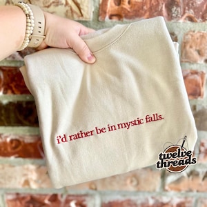 May include: A beige crewneck sweatshirt featuring the embroidered red text "i'd rather be in mystic falls." centered on the chest. The garment has a ribbed collar and long sleeves, with a small circular logo in the bottom right corner.