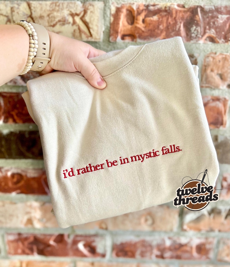 May include: A beige crewneck sweatshirt featuring the embroidered red text "i'd rather be in mystic falls." centered on the chest. The garment has a ribbed collar and long sleeves, with a small circular logo in the bottom right corner.