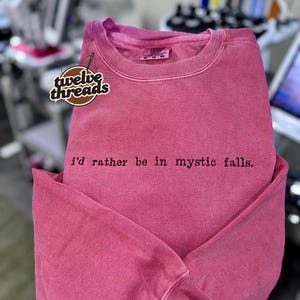 May include: A faded pink crewneck sweatshirt featuring the black embroidered text "i'd rather be in mystic falls." on the chest. The garment includes a branded neck label and a logo patch reading "twelve threads" on the upper left side.