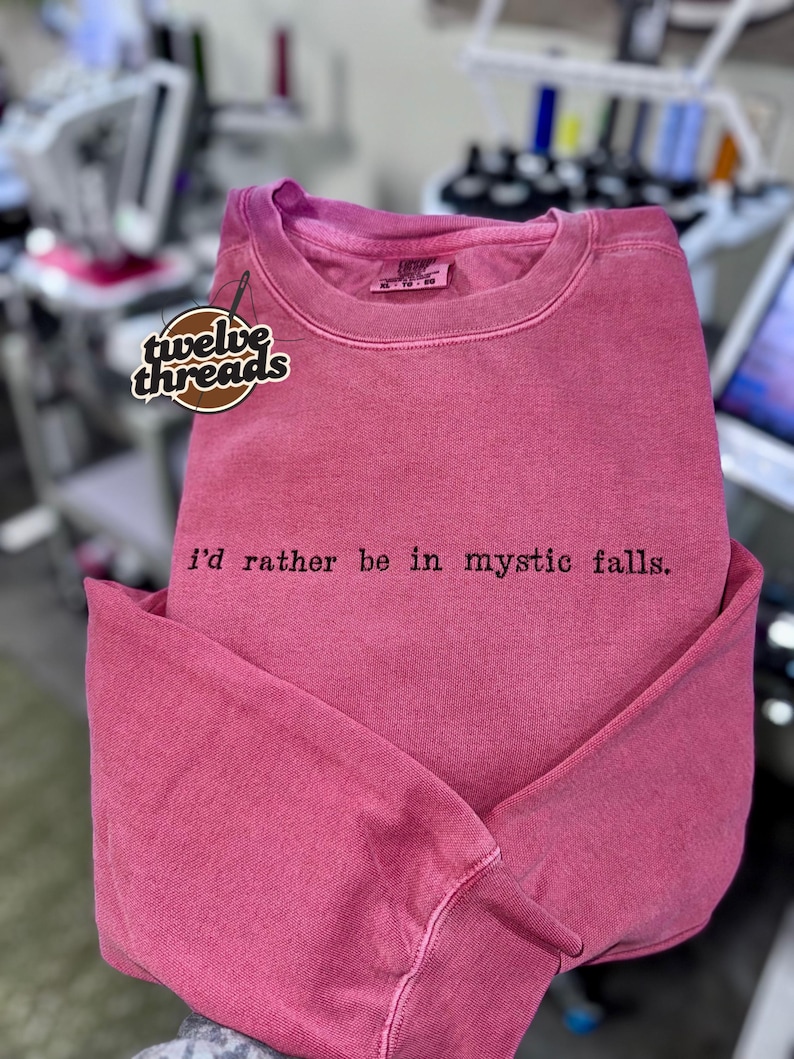 May include: A faded pink crewneck sweatshirt featuring the black embroidered text "i'd rather be in mystic falls." on the chest. The garment includes a branded neck label and a logo patch reading "twelve threads" on the upper left side.