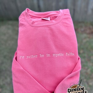 May include: A bright pink crewneck sweatshirt featuring the white embroidered text "i'd rather be in mystic falls." across the chest. The garment has a ribbed collar, long sleeves, and a visible brand tag inside the neckline.