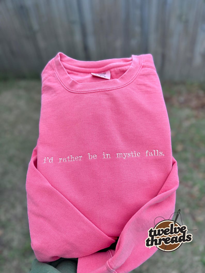 May include: A bright pink crewneck sweatshirt featuring the white embroidered text "i'd rather be in mystic falls." across the chest. The garment has a ribbed collar, long sleeves, and a visible brand tag inside the neckline.