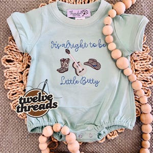 May include: A light blue baby romper with the text "It's alright to be Little Bitty" and embroidered images of cowboy boots, a guitar, and a cowboy hat. The romper has short sleeves and a round neckline. The brand logo "twelve threads" is also visible.