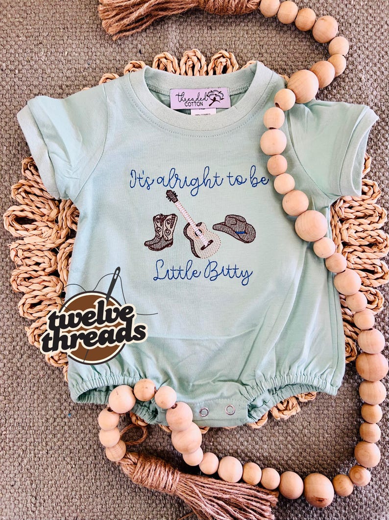 May include: A light blue baby romper with the text "It's alright to be Little Bitty" and embroidered images of cowboy boots, a guitar, and a cowboy hat. The romper has short sleeves and a round neckline. The brand logo "twelve threads" is also visible.