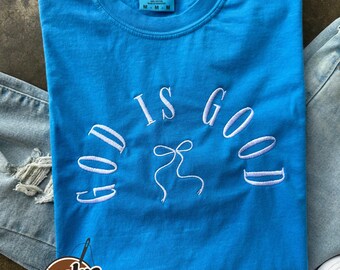 Puede incluir: Una camiseta turquesa Comfort Colors con la frase bordada "GOD IS GOOD" en escritura blanca, con un diseño de lazo. La camiseta está colocada sobre unos vaqueros, con zapatillas blancas visibles.