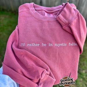 May include: A dusty rose long-sleeve crewneck sweatshirt featuring the embroidered white text "i'd rather be in mystic falls." across the chest. The garment has a ribbed collar, cuffs, and hem with a faded, vintage-inspired fabric finish.