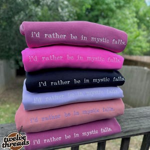 May include: A stack of six folded sweatshirts in various colors including mauve, bright pink, black, light purple, dusty rose, and dark magenta. Each sweatshirt features the embroidered white text "i'd rather be in mystic falls."