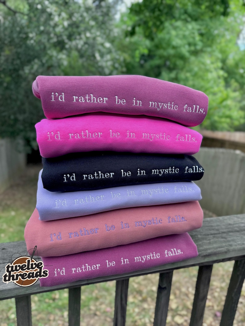 May include: A stack of six folded sweatshirts in various colors including mauve, bright pink, black, light purple, dusty rose, and dark magenta. Each sweatshirt features the embroidered white text "i'd rather be in mystic falls."