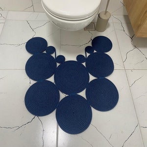 May include: A dark blue bathroom rug made of braided rope, shaped like a stylized heart with multiple circles. The rug is on a white tiled floor.