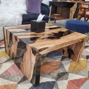 May include: A wooden coffee table with a black epoxy resin top. The table has a natural wood grain and a unique, organic shape. The table is sitting on a colourful rug with geometric patterns.