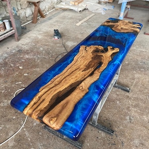 Puede incluir: Una tapa de mesa larga y rectangular hecha de madera y resina epoxi azul. La madera tiene un patrón de veta natural y la resina epoxi tiene un efecto de remolino y mármol. La tapa de la mesa está soportada por dos patas de metal.