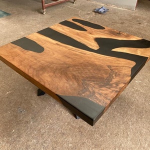 May include: A square coffee table with a wooden top and black epoxy resin inlay. The table has a natural wood grain pattern and a modern design.