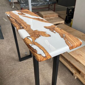 Könnte beinhalten: Ein langer, schmaler Konsolentisch mit einer weißen Epoxidharzplatte und einem Untergestell aus Naturholz. Der Tisch hat ein modernes Design und wird von zwei schwarzen Metallbeinen getragen.